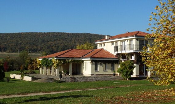 Két szintes családi ház, piros cserépfedéllel és terasszal, zöld környezetben.