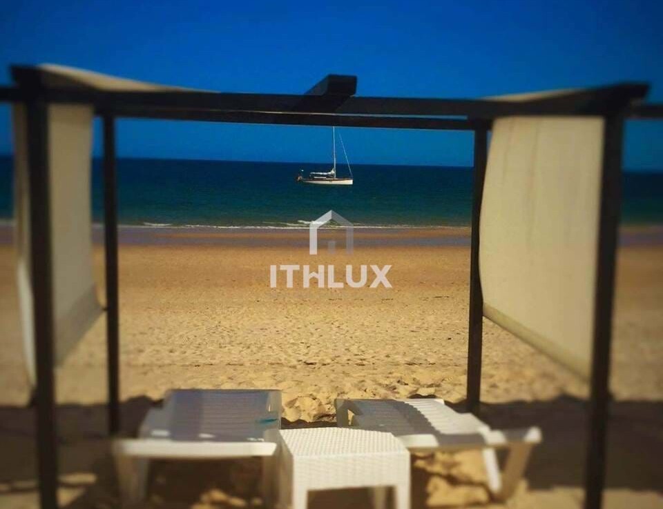 Fehér napernyő és két fehér napozóágy a tengerparton, háttérben a tengerrel.