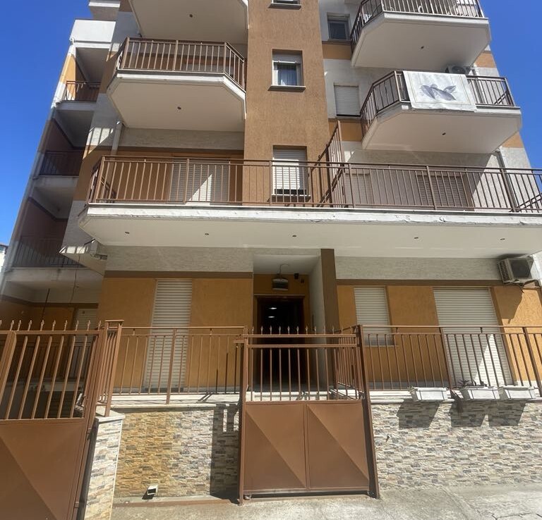 Többemeletes lakóépület bejárata, erkélyekkel és kerítéssel.