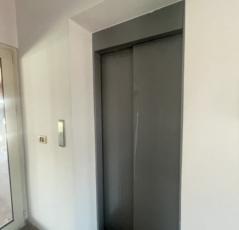 Szürke lift ajtó egy épület falában, mellette lift hívó gomb.